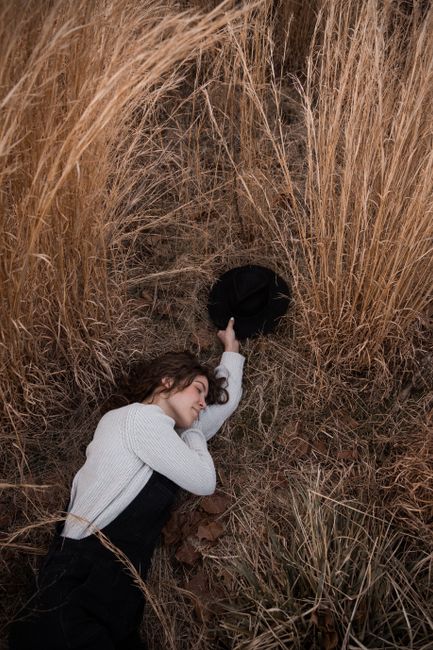 A woman peacefully lies in tall dry grass, holding a black hat, capturing a serene outdoor moment.