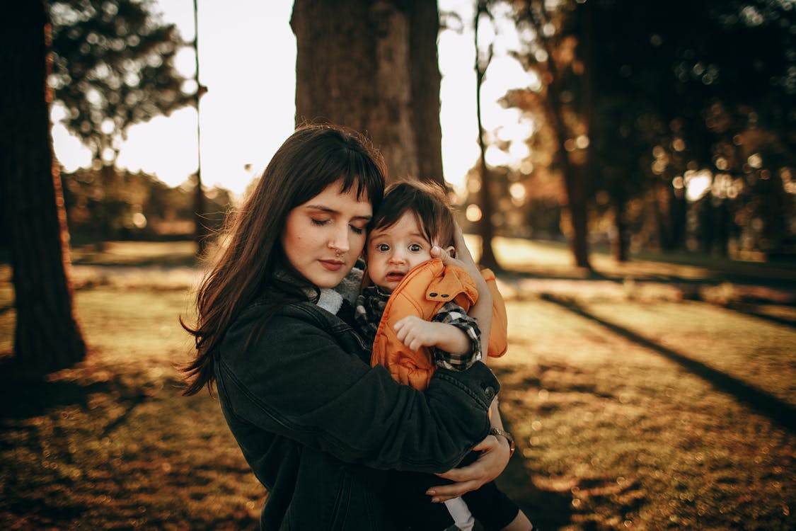 Loving mother hugging cute little son in warm clothes while standing near tree in park in sunny summer evening