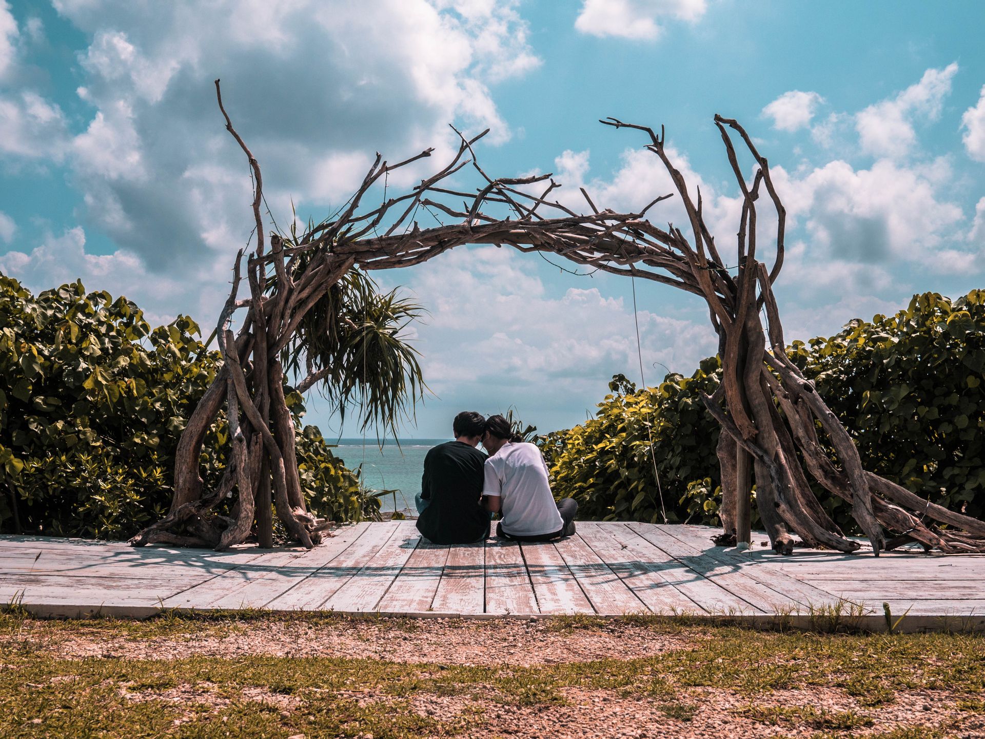 A couple sits peacefully under a driftwood arch on a sunny beach day, surrounded by serene nature.
