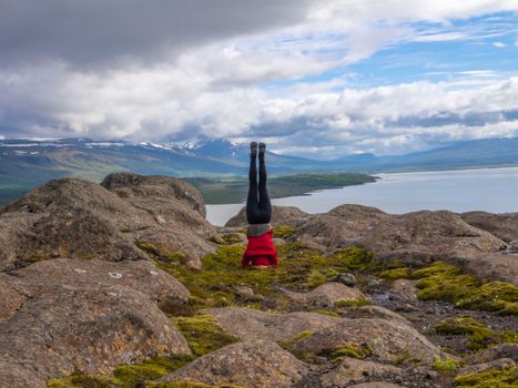 A person in a red jacket performs a headstand on a rocky mountain landscape with a lake view.