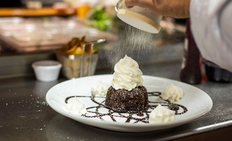 A spoon drizzling white powdered sugar icing over a batch of glazed cookies, with a bowl of icing and a small pitcher of milk beside it.