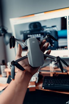 A close-up view of a man's hand holding a drone in front of a computer screen displaying camera equipment.