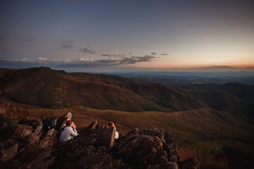 Free From above of unrecognizable travelers sitting on rocky hill and enjoying scenic view of mountains in evening Stock Photo