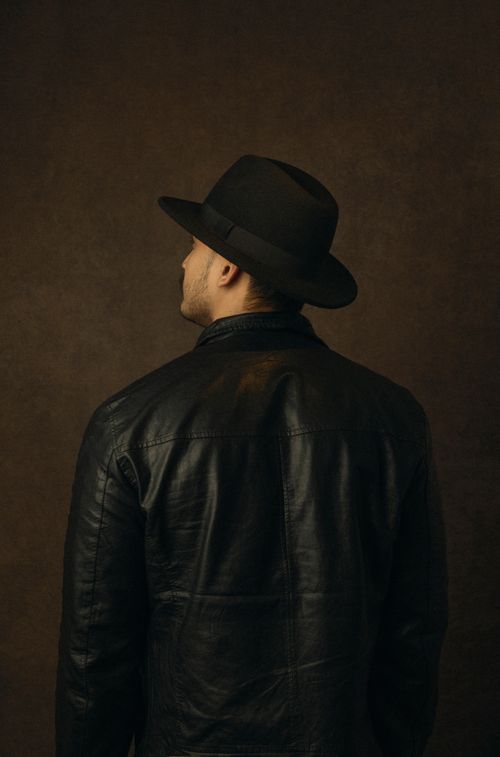 Free Rear view of a man wearing a fedora and leather jacket, set against a dark background. Stock Photo