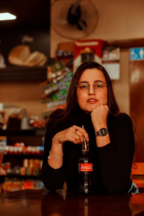 Free Portrait of a woman with eyeglasses sitting indoors, holding a Coca-Cola, in a warm and cozy cafe setting. Stock Photo