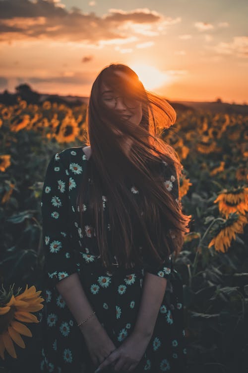 Free A Woman in Black and White Floral Dress Standing on Sunflower Field during Sunset Stock Photo