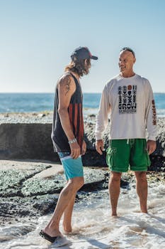Two men smiling and standing on a rocky shore, enjoying a sunny day by the water.
