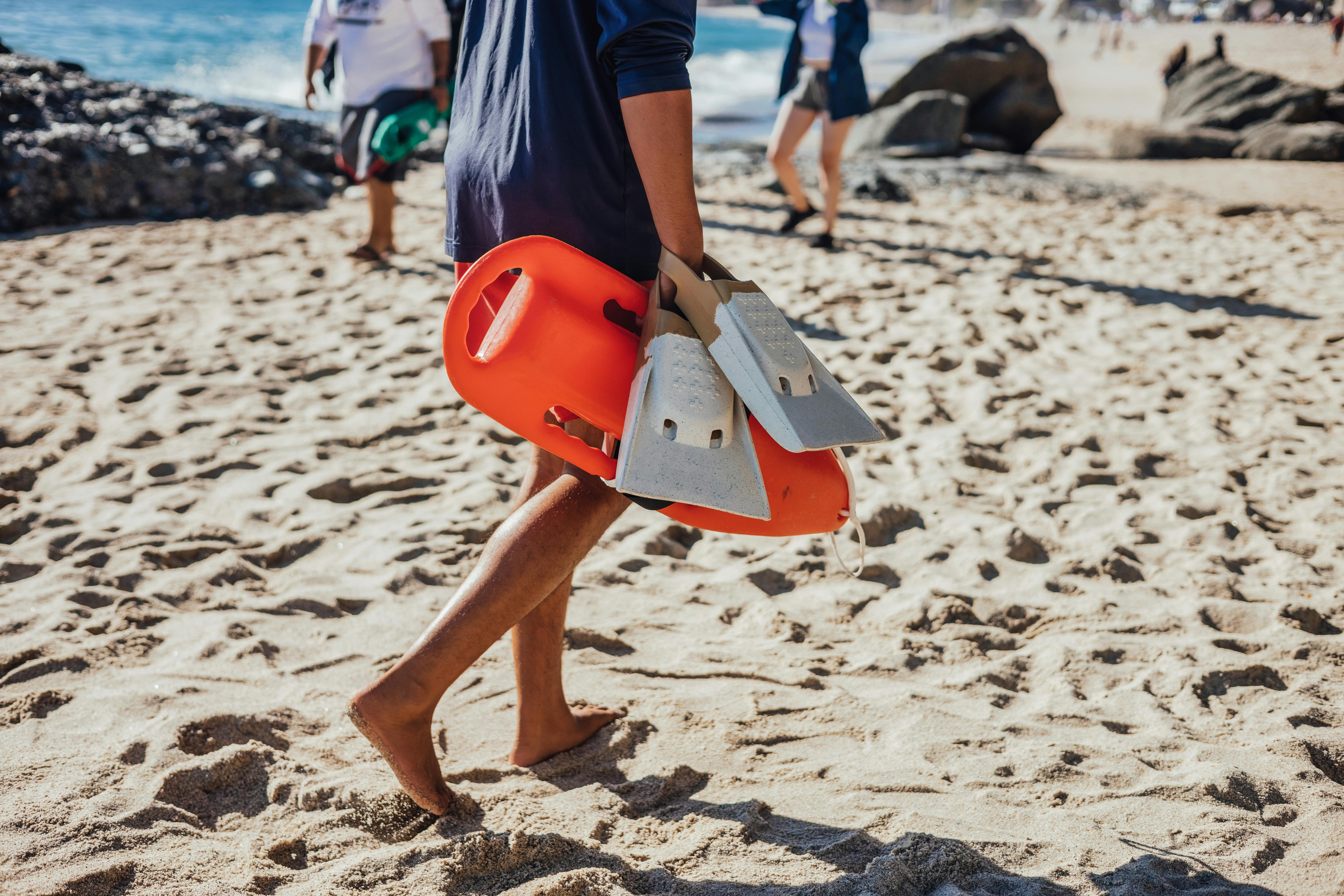 Lifeguard carrying rescue equipment and swim fins while walking on a sunny beach day.