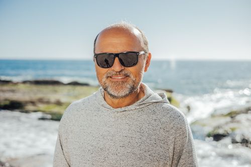Free Senior man wearing sunglasses, smiling at the beach on a sunny day. Stock Photo