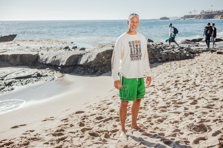 An Elderly Man In White Sweatshirt Smiling While Standing On The Beach Sand