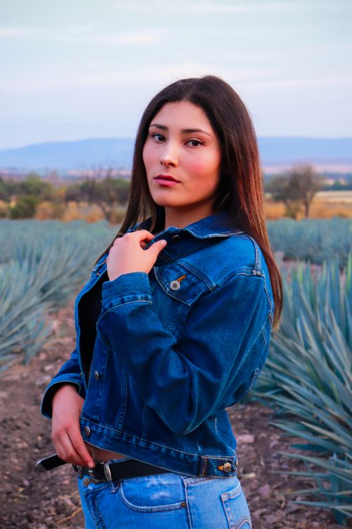 Free Portrait of a young woman in a denim jacket standing on a dirt road surrounded by plants at sunset. Stock Photo