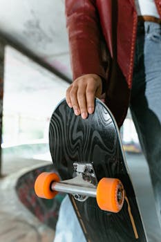 Hand gripping skateboard deck in urban skate park, wearing red leather jacket.