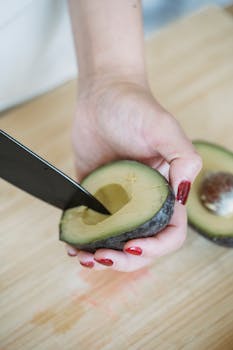 Close-up of a hand slicing an avocado on a wooden board with a knife.