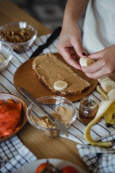 Close-up of hands preparing a banana peanut butter sandwich with fresh ingredients on a wooden board.