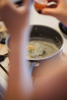 Close-up of a poached egg cooking in a pot on a gas stove, hands in view.