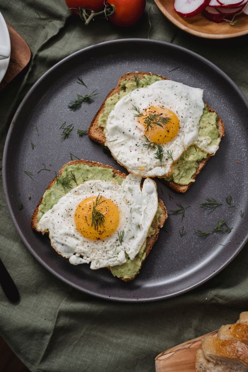 Free Delicious avocado toast topped with fried eggs on a rustic dark plate, perfect for a hearty breakfast. Stock Photo