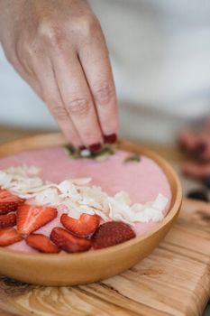Close-up of a hand garnishing a smoothie bowl with strawberries and coconut flakes.