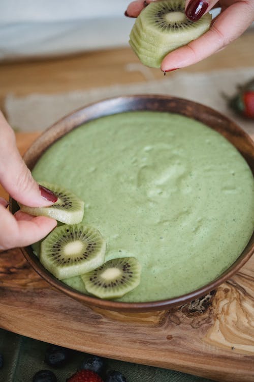 Person Putting Slices of Kiwi on Top of a Green Smoothie