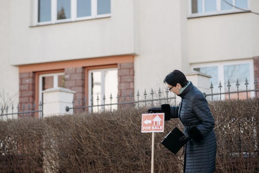 Real estate agent in a black coat placing a 'For Sale' sign in front of a house.