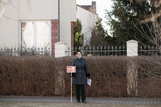 Real estate agent outside a house holding a 'For Sale' sign in winter.