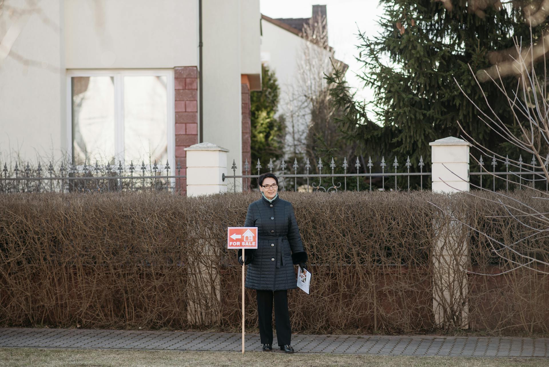 Woman in Jacket Standing by House and Holding For Sale Banner