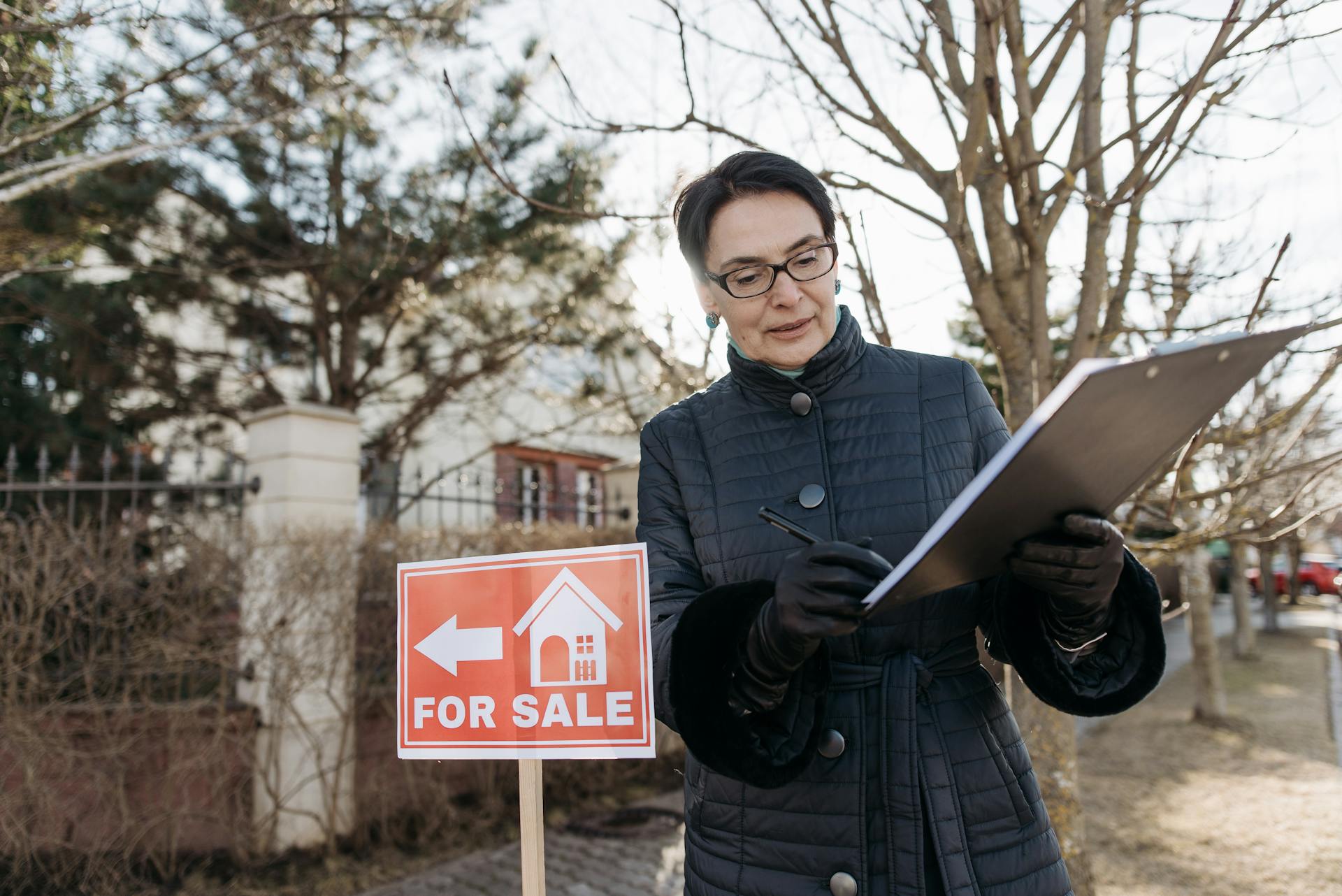 A real estate agent reviews a clipboard next to a 'For Sale' sign outside a property on a clear day.