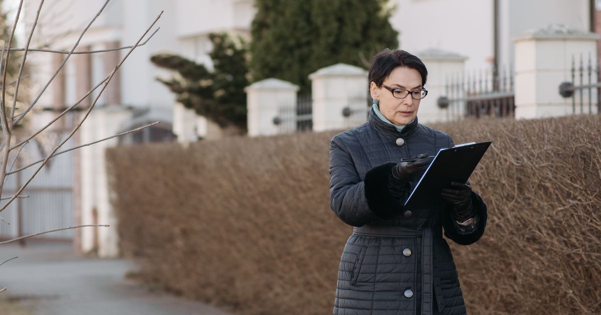 A property inspector with a clipboard reviews credentials and notes outside a suburban residential home, representing the on-site side of contractor background checks.