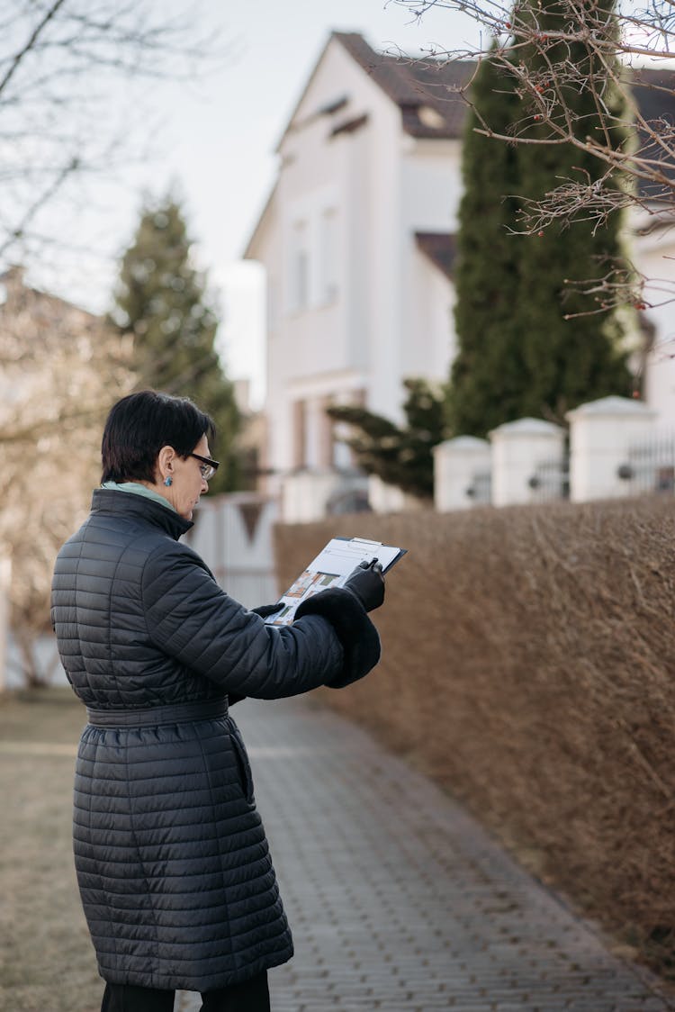 Senior Woman In Black Winter Jacket Reading A Document While Standing On A Walkway