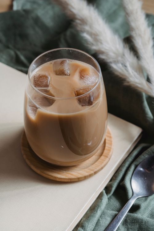 Free A close-up of iced coffee in clear glass with ice cubes on a wooden coaster, surrounded by pampas grass. Stock Photo