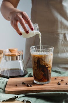 A person pouring milk into a glass of cold brew coffee on a wooden tray.