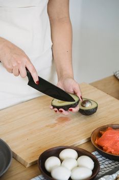 A person slicing an avocado on a wooden cutting board with eggs and salmon nearby.