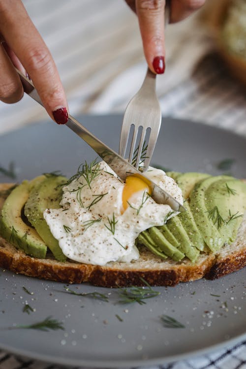 Slicing Poached Egg on Top of Toasted Bread with Green Fresh Fruit and Rosemary