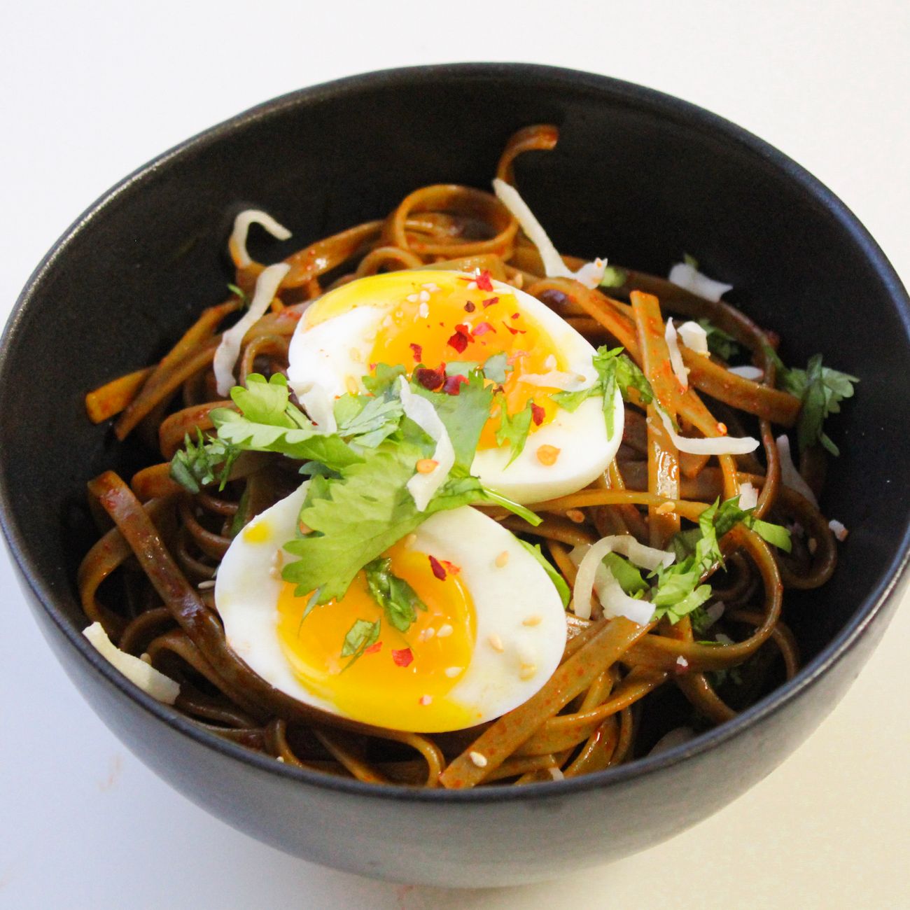 Bowl of dark buckwheat noodles topped with julienned vegetables, halved soft-boiled egg, and red gochujang sauce, garnished with sesame seeds and scallions