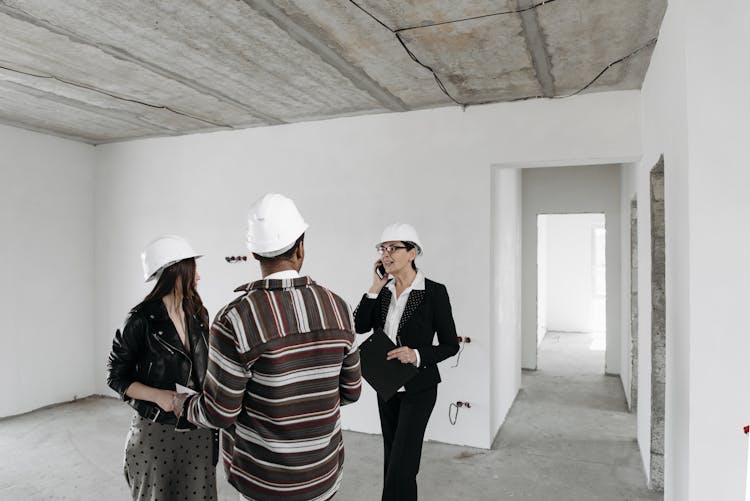 Real Estate Agent And Young Couple In Helmets Standing In An Empty Room With White Walls