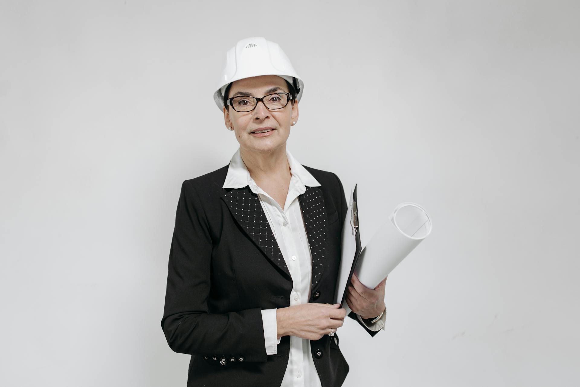 Confident female architect in a suit and hard hat holding blueprints against a white background.