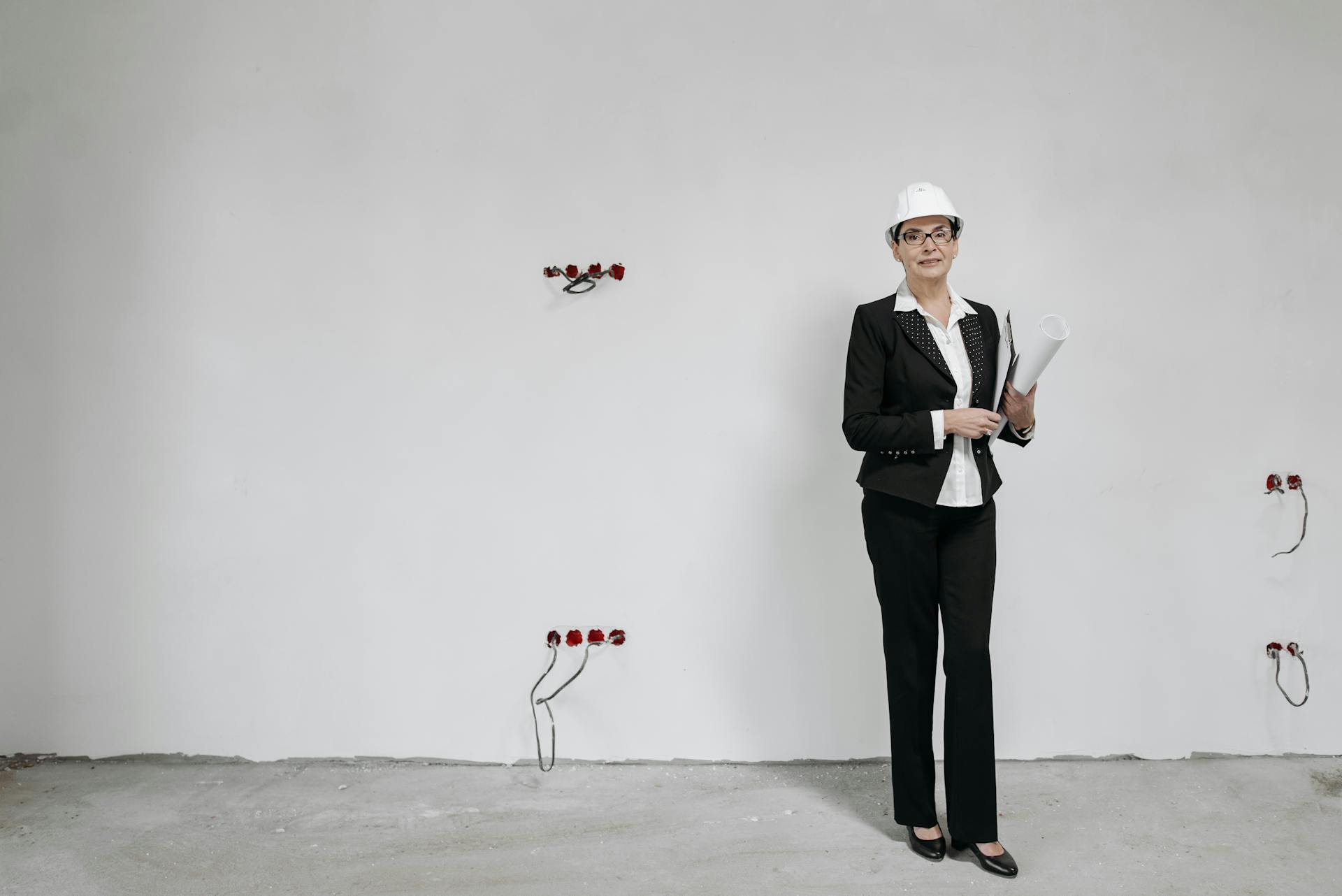 Woman architect in suit holding blueprints, standing by a white wall in an unfinished space.