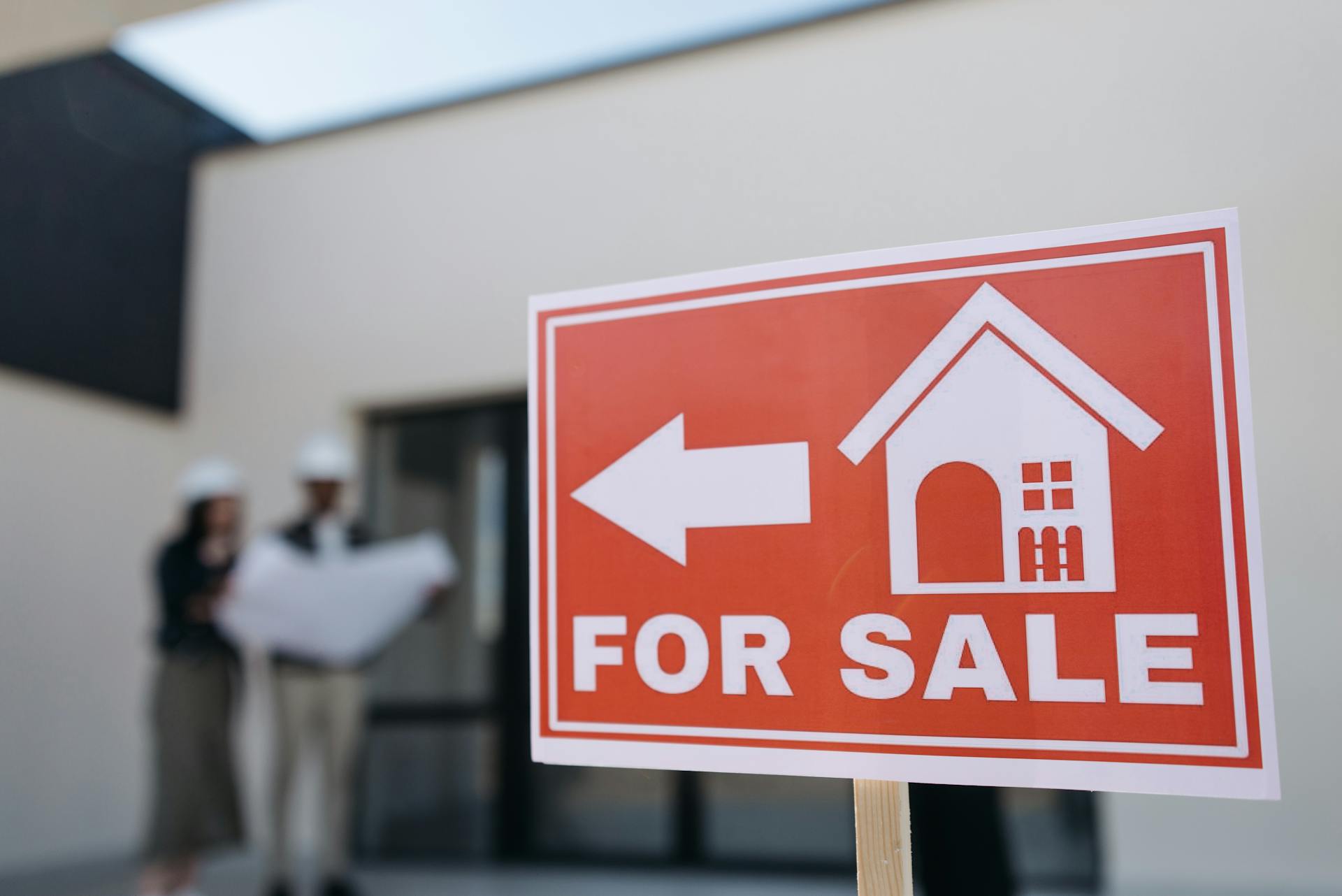 A real estate sign indicates a property for sale as two agents in hard hats discuss building plans outdoors.