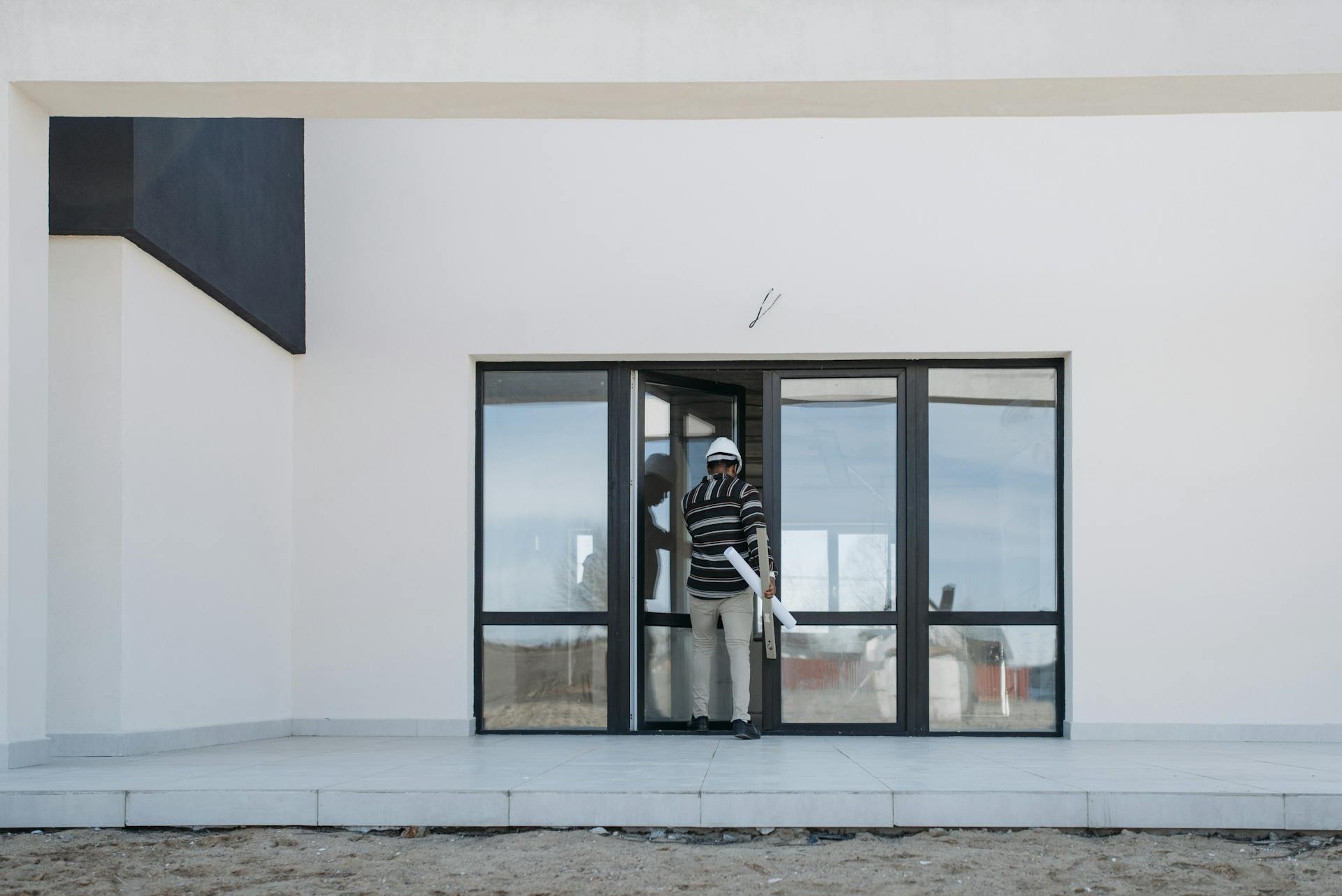 Architect wearing helmet and holding blueprints exits a modern glass building.