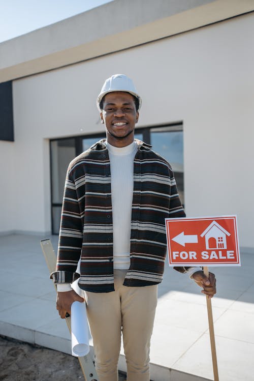 Man Wearing Hard Hat Holding a For Sale Sign while Smiling at the Camera