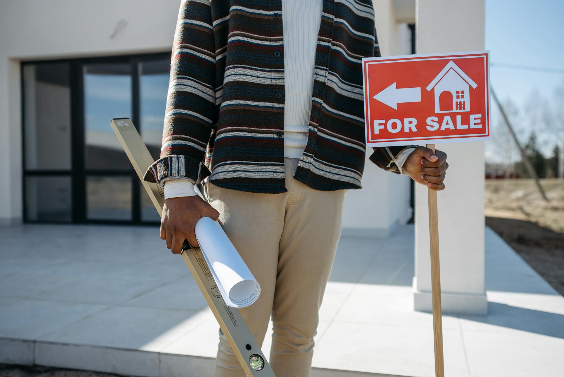 A real estate agent stands outside with a 'for sale' sign and rolled blueprint, suggesting a property for sale.