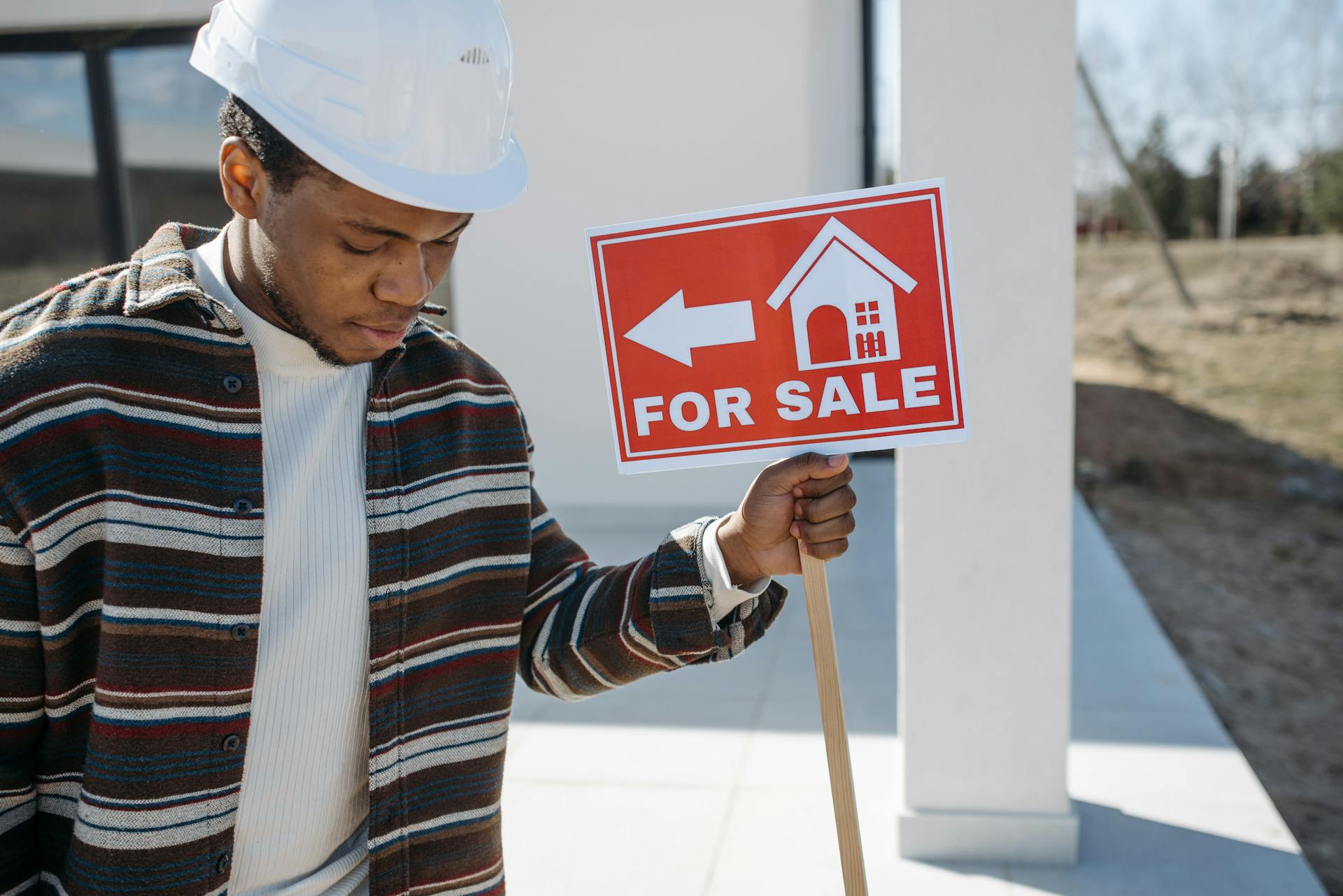 Young man with a hard hat holding a red for sale sign outside a building.