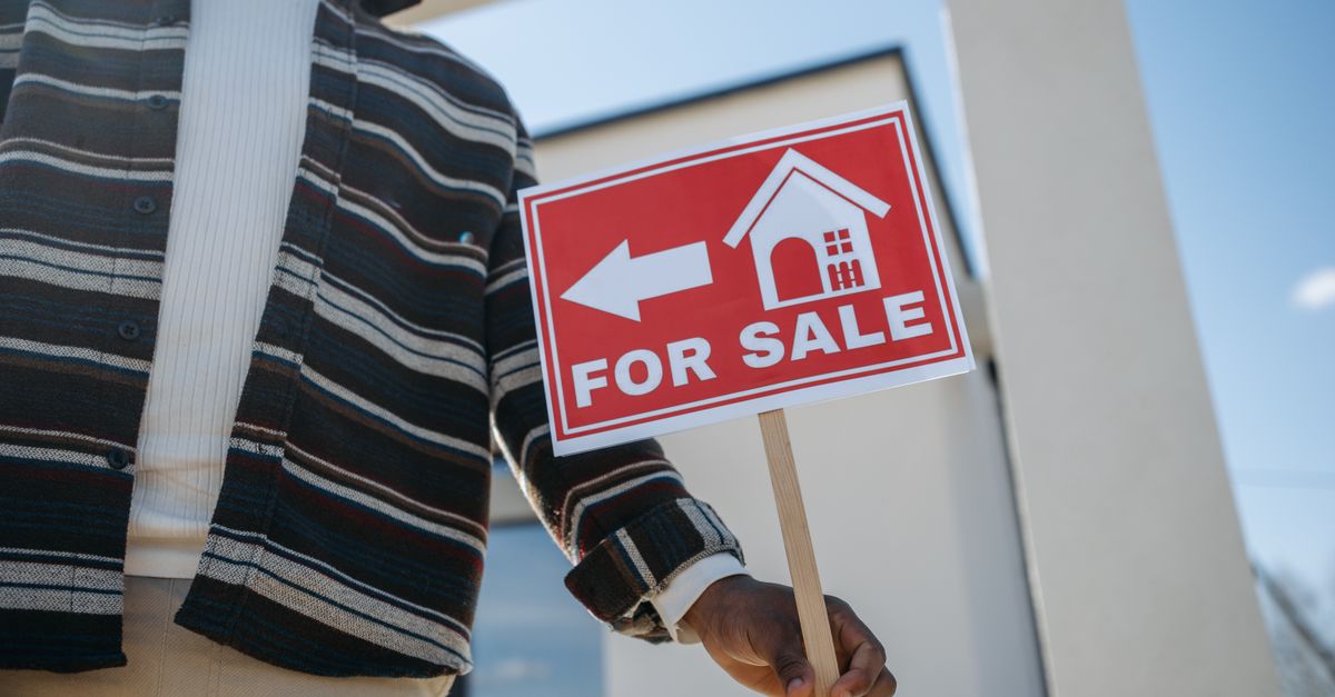 Close-up of a person holding a for sale sign outside a modern house.