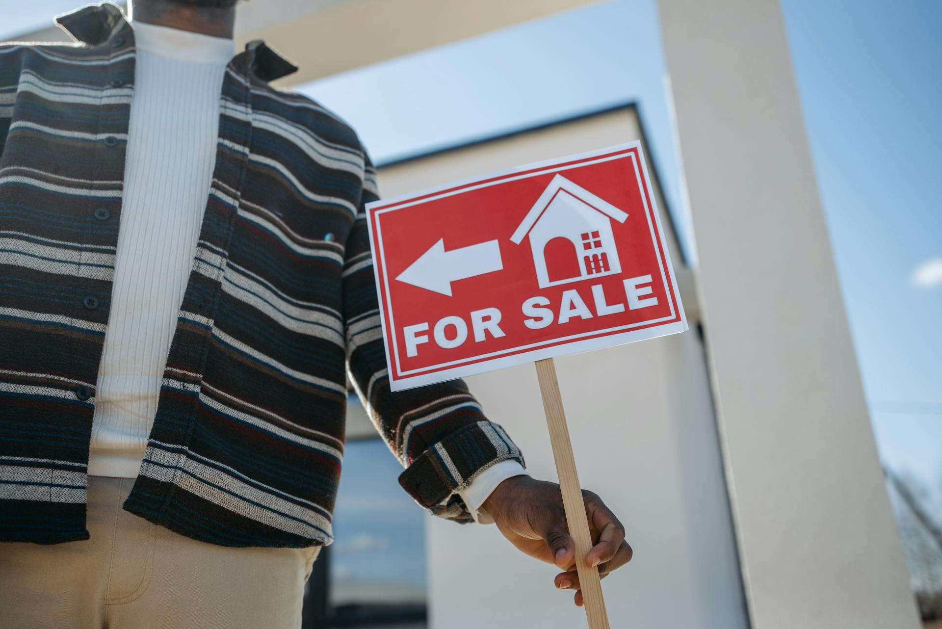 Close-up of a person holding a for sale sign outside a modern house.