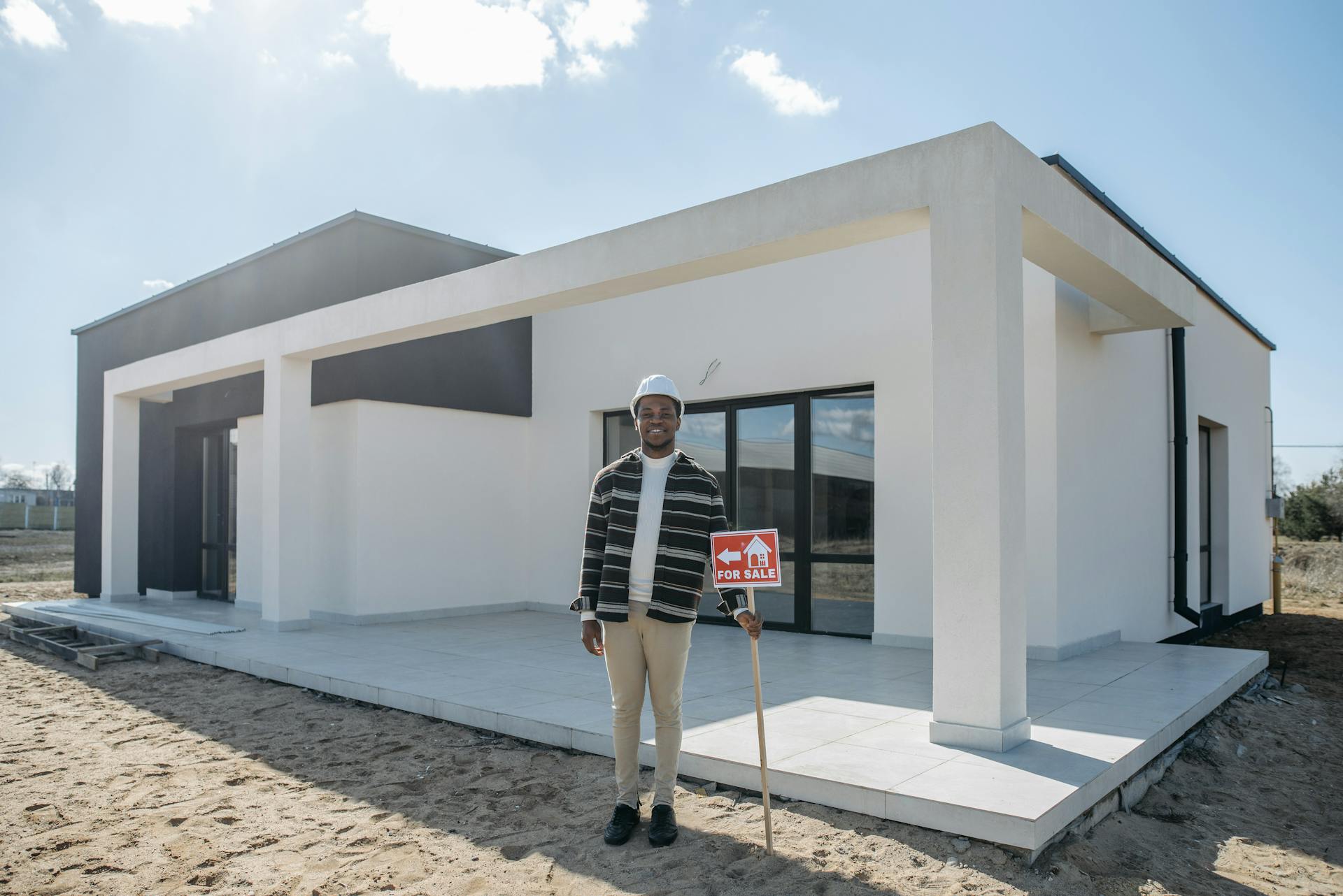 A realtor with a hard hat holds a for sale sign in front of a modern house, ready for sale.