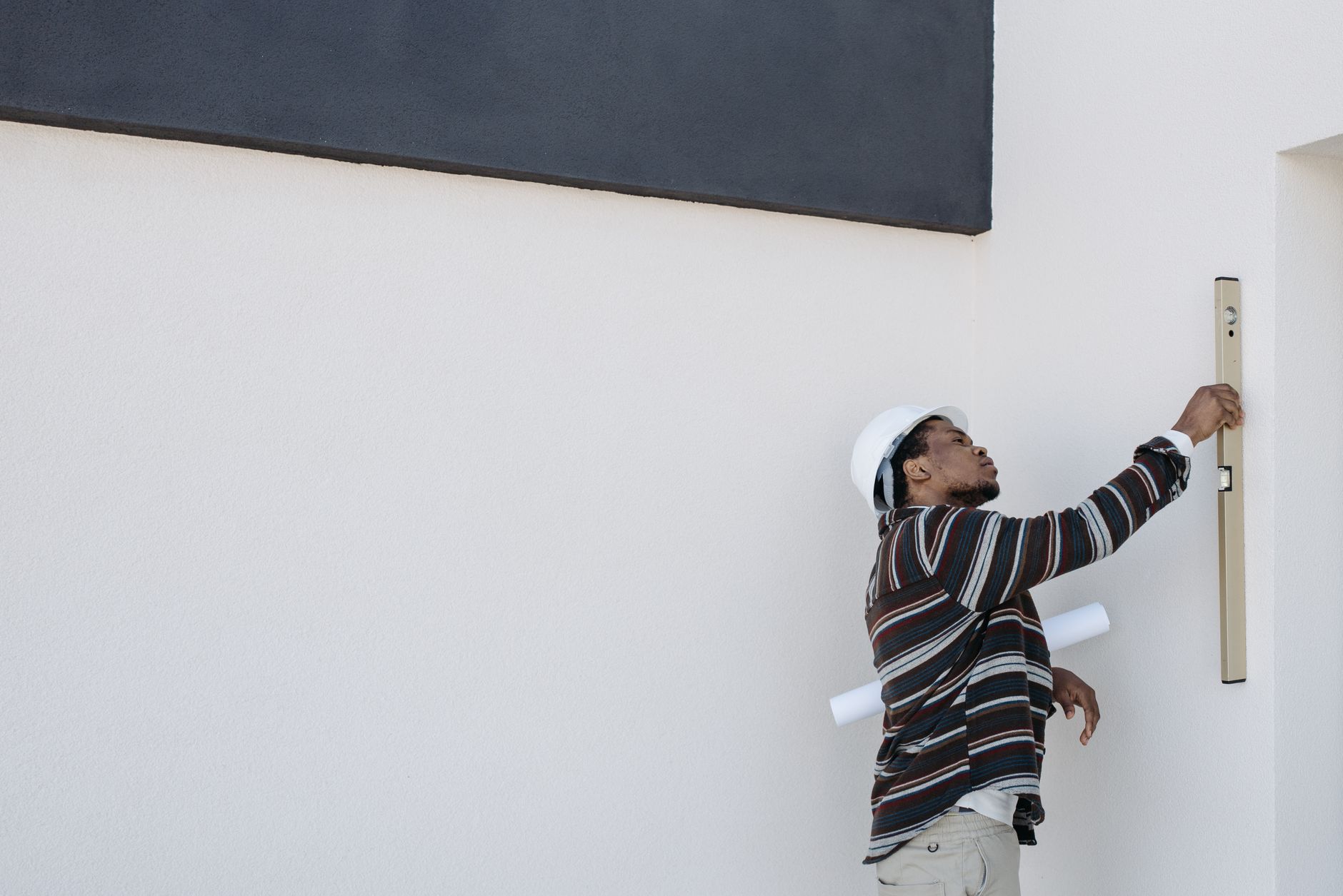 An architect wearing a hard hat checks wall alignment with a level tool outdoors.