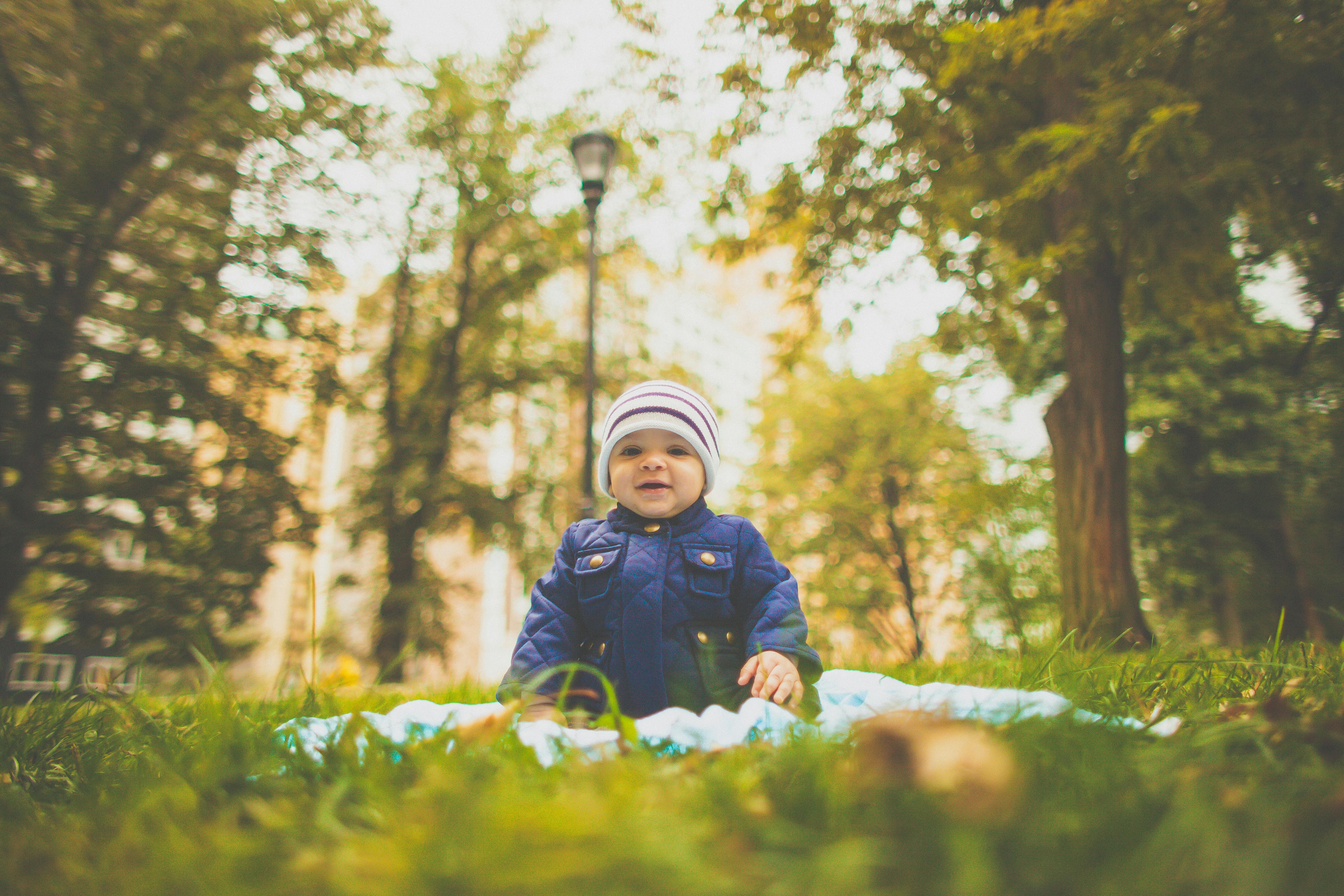 Cute baby sitting on a picnic blanket in a lush green park wearing winter clothing.