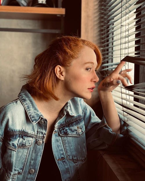 Free A Woman in Denim Jacket Looking Through Window Blinds Stock Photo