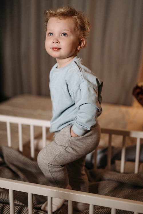 Toddler in Blue Long Sleeve Shirt and Gray Pants Standing on Baby Bed