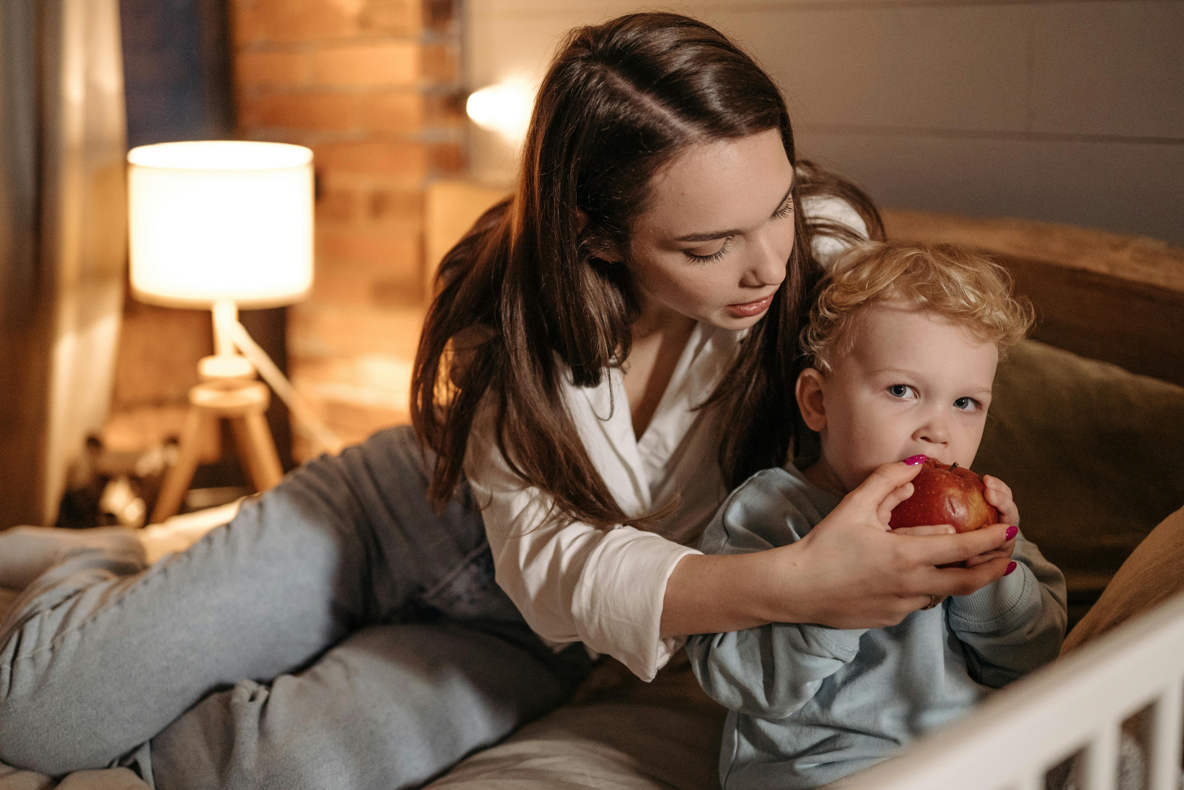 Warm scene of a mother assisting her young child with eating an apple in a cozy bedroom setting.