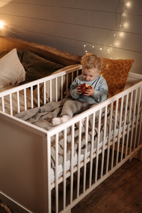 A Cute Baby Sitting on the Crib while Holding an Apple